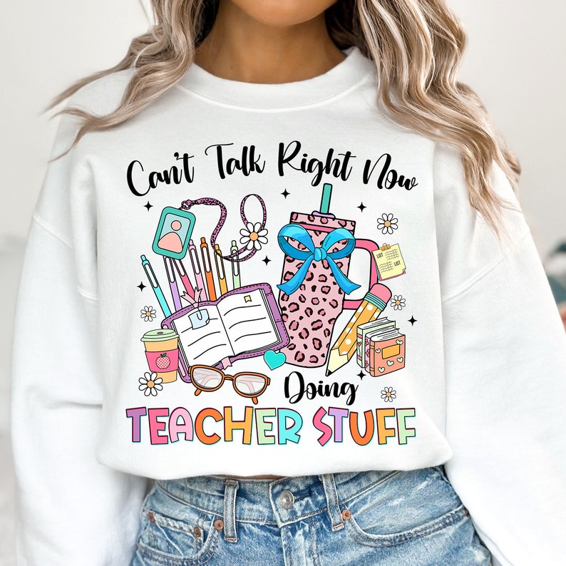 May include: White sweatshirt with a graphic design of school supplies and the text "Can't Talk Right Now Doing Teacher Stuff".