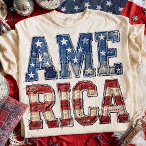 May include: Cream-colored t-shirt with the word "AMERICA" in patchwork denim and red fabric. The letters are outlined with stitching and feature white stars. A patriotic design.