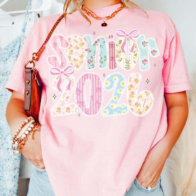 Op de afbeelding: Roze t-shirt met de tekst "Senior 2026" in een bloemen- en lintontwerp. Het shirt heeft een relaxte pasvorm en korte mouwen. De tekst is in patchwork-stijl met diverse bloempatronen en strikken.