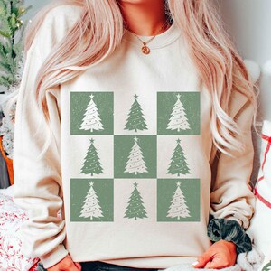 May include: A cream-colored sweatshirt with a pattern of nine green and white Christmas trees. The trees are arranged in a grid pattern.
