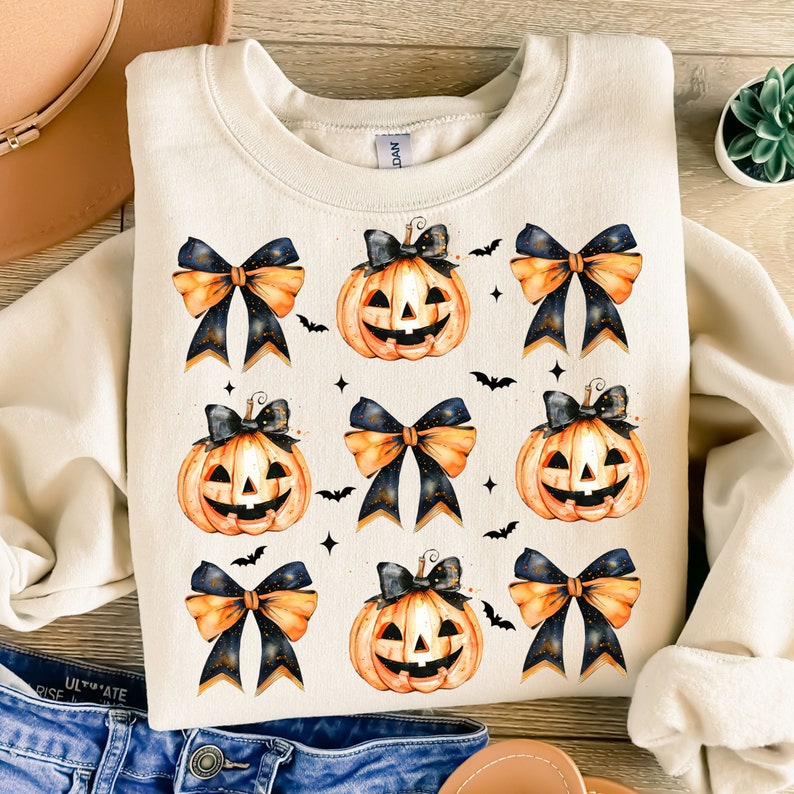 May include: A cream-colored sweatshirt with a repeating pattern of orange pumpkins with black eyes and a toothy grin, each pumpkin topped with a black and orange bow. The background is white with small black bats and gold glitter.