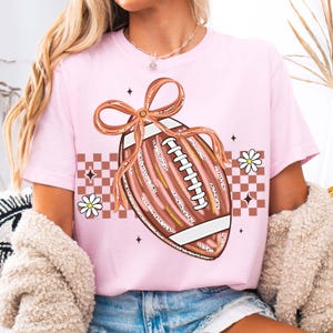 May include: A light pink t-shirt featuring a football design with a bow, daisies, and a checkered pattern. The football is brown with white laces and gold accents. The design is centered on the shirt.