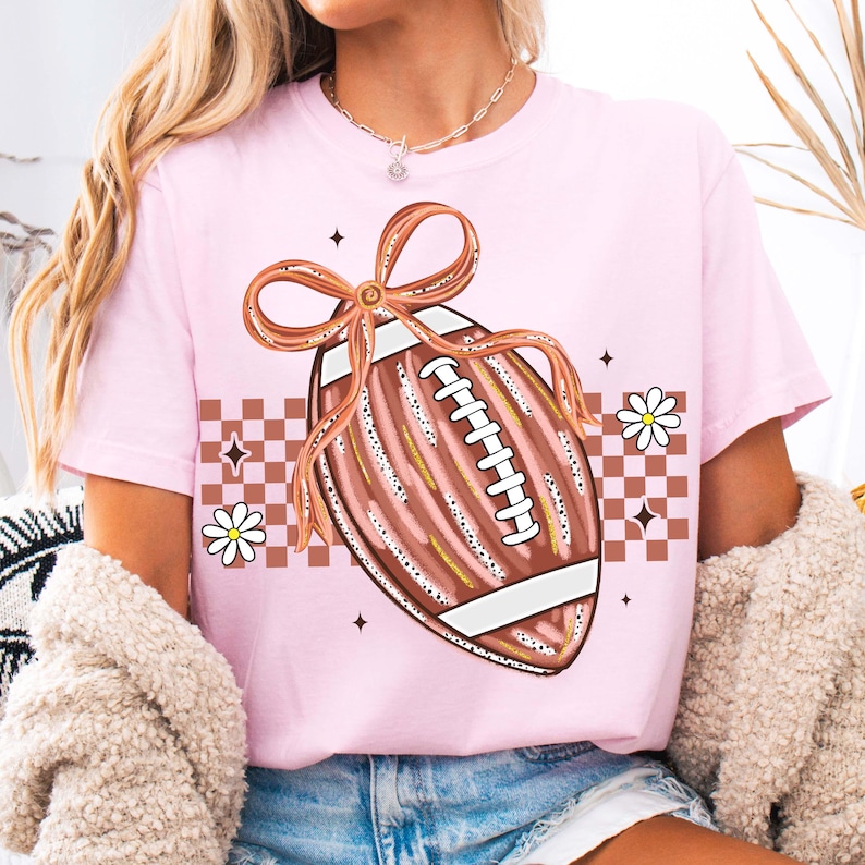 May include: A light pink t-shirt featuring a football design with a bow, daisies, and a checkered pattern. The football is brown with white laces and gold accents. The design is centered on the shirt.