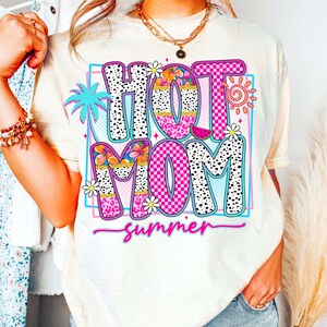Puede incluir: Camiseta color crema con un gr&aacute;fico colorido que dice "HOT MOM summer". El dise&ntilde;o incluye una palmera, flores, un sol y una rodaja de sand&iacute;a. El texto utiliza una mezcla de fuentes y patrones, incluyendo lunares y cuadros.
