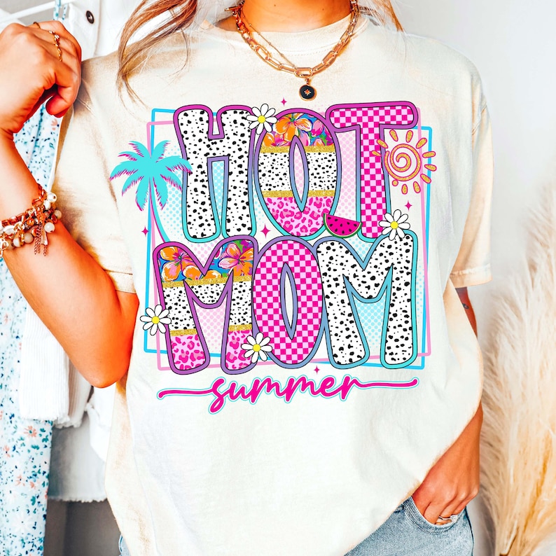 Puede incluir: Camiseta color crema con un gr&aacute;fico colorido que dice "HOT MOM summer". El dise&ntilde;o incluye una palmera, flores, un sol y una rodaja de sand&iacute;a. El texto utiliza una mezcla de fuentes y patrones, incluyendo lunares y cuadros.