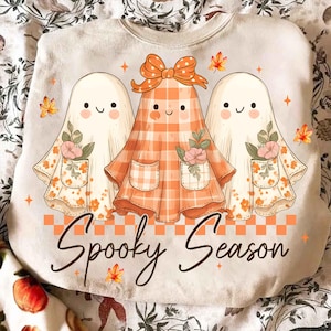 May include: A cream-colored sweatshirt featuring three cartoon ghosts. The central ghost wears an orange plaid dress with a bow, while the others have floral accents. The phrase "Spooky Season" is written in a script font.