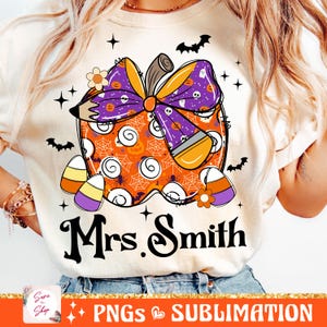 Pode incluir: Uma camiseta branca com um desenho de abóbora laranja, roxa e preta. A abóbora é decorada com milho doce, um laço e um lápis. O texto "Mrs. Smith" é impresso abaixo da abóbora.