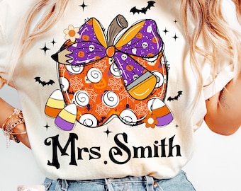 May include: A white t-shirt featuring an orange, purple and black pumpkin design. The pumpkin is decorated with candy corn, a bow and a pencil. The text "Mrs. Smith" is printed below the pumpkin.