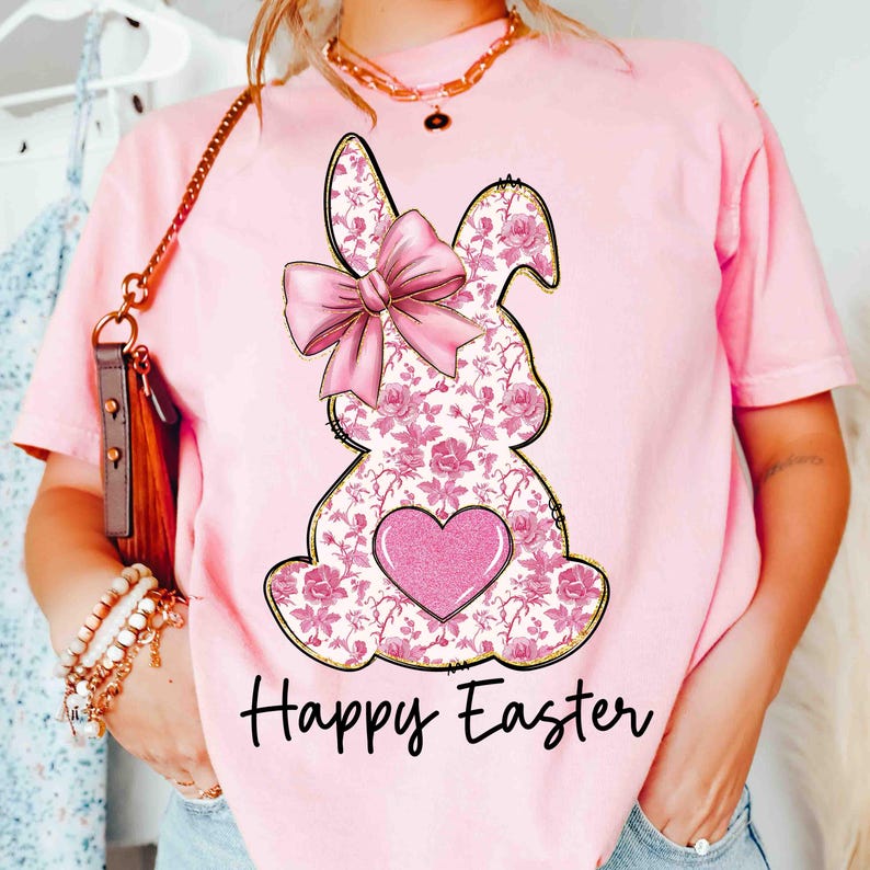 Op de afbeelding: Roze T-shirt met een bloemig konijnenontwerp en een roze strik. Het konijn heeft een roze hart in het midden. De tekst "Happy Easter" is onder het konijn gedrukt.