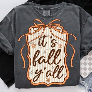 May include: Dark gray sweatshirt with a decorative orange and white graphic that reads "it's fall y'all". The graphic has a bow and floral accents. The sweatshirt is a comfortable, casual style.