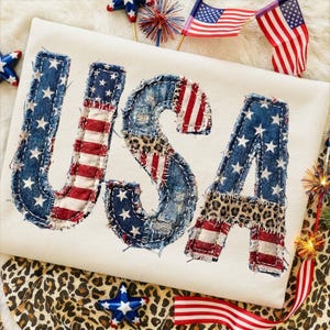 Pu&ograve; includere: Una maglietta color crema con la scritta "USA" applicata in denim, strisce della bandiera americana e stampa leopardata. Le lettere sono cucite con filo bianco, un design patriottico.