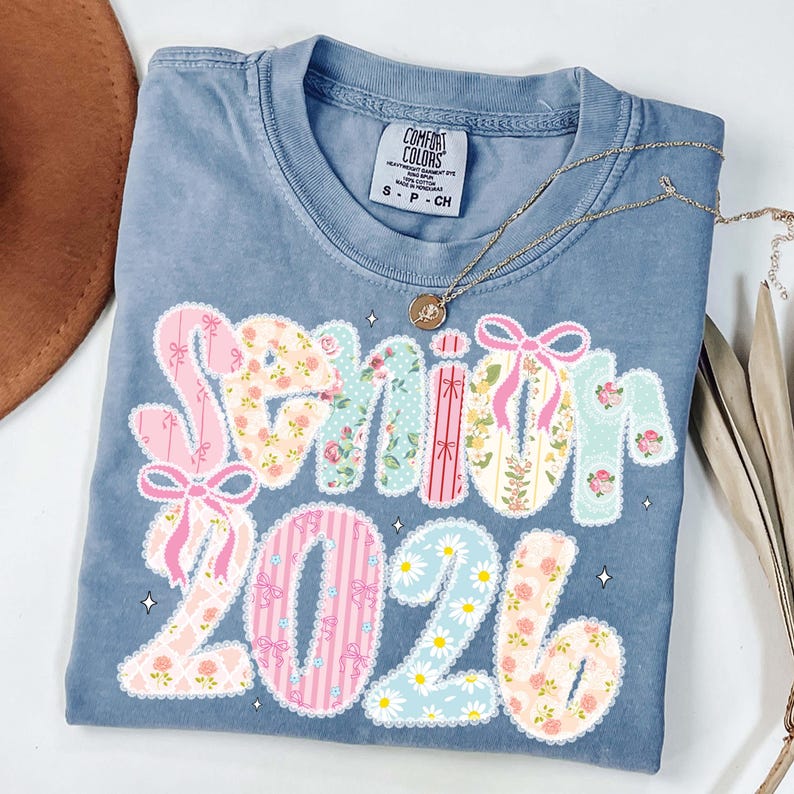 Op de afbeelding: Een blauw Comfort Colors T-shirt met de tekst "Senior 2026" in een patchwork-ontwerp. Het woord "Senior" is in bloemenpatronen en "2026" in madeliefjespatronen. Een gouden ketting ligt over het shirt.