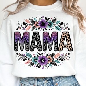 May include: White sweatshirt featuring a purple, pink and black "MAMA" graphic with a leopard print design. The graphic is surrounded by floral designs with pink, purple and teal flowers.