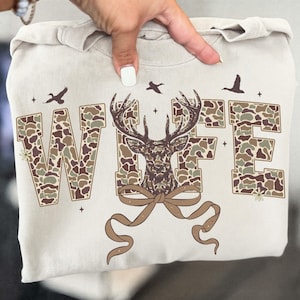 May include: A beige sweatshirt featuring the word "WIFE" in camouflage print, with a deer graphic in the center. The design includes a brown bow, birds, and small floral accents. The sweatshirt is held up by a hand.