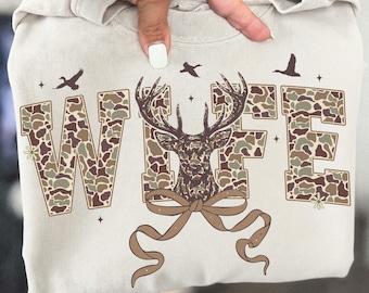 May include: A beige sweatshirt featuring the word "WIFE" in camouflage print, with a deer graphic in the center. The design includes a brown bow, birds, and small floral accents. The sweatshirt is held up by a hand.