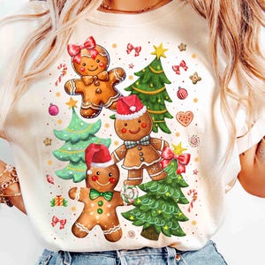 Könnte beinhalten: Ein weißes T-Shirt mit einem Weihnachts-Lebkuchen-Motiv. Das Design zeigt drei Lebkuchenmänner, zwei Weihnachtsbäume und verschiedene festliche Dekorationen wie Zuckerstangen, Schleifen und Ornamente.