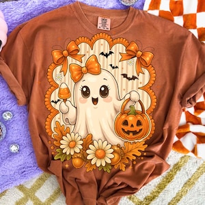 Pode incluir: Camiseta cor de ferrugem com um fantasma de desenho animado fofo segurando uma ab&oacute;bora e doces de milho. O fantasma &eacute; cercado por la&ccedil;os laranja, morcegos e flores, com uma borda decorativa. A camisa tem decote redondo e mangas curtas.