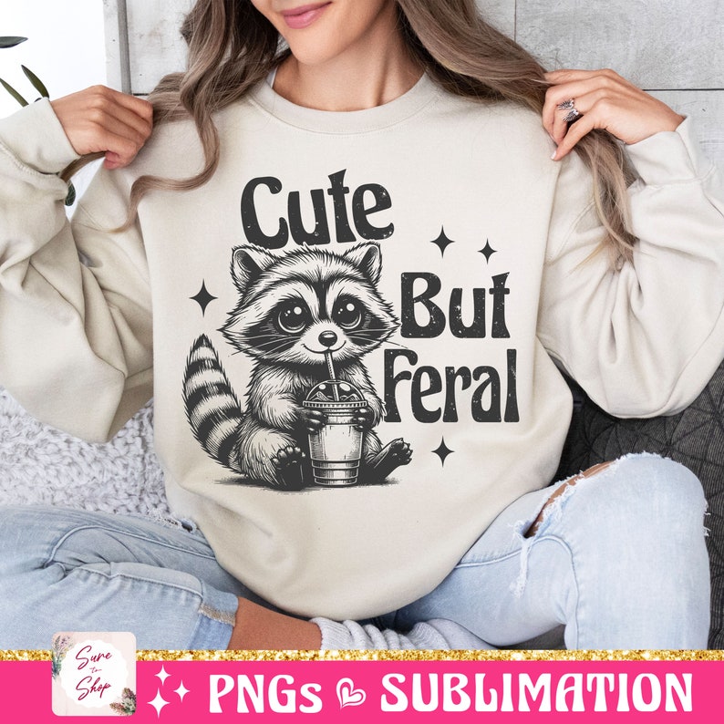 May include: A cream-colored sweatshirt featuring a raccoon illustration holding a drink with the text "Cute But Feral". The raccoon is black and white with large eyes. The design includes star accents.