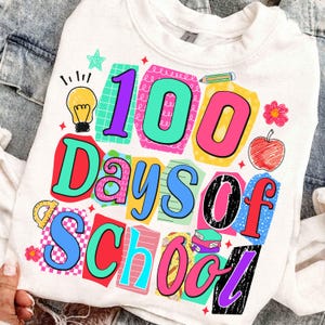 Puede incluir: Sudadera blanca con un gr&aacute;fico colorido que dice "100 Days of School". El dise&ntilde;o incluye una bombilla, una estrella, una manzana y &uacute;tiles escolares. El texto es de varios colores brillantes.