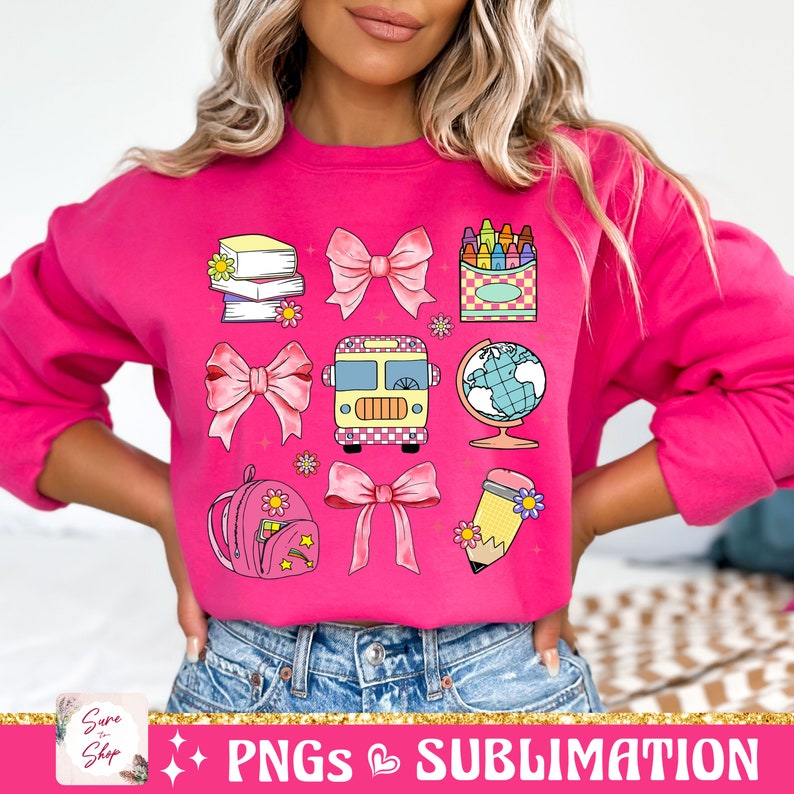 May include: Pink sweatshirt with a colorful back-to-school design featuring a school bus, books, a globe, a pencil, a backpack, crayons, and bows.