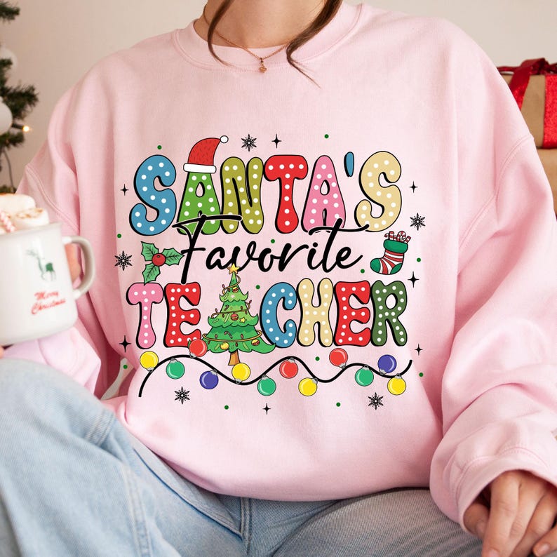 May include: Pink sweatshirt with a colorful graphic that says "Santa's Favorite Teacher" with a Christmas tree, ornaments, and a stocking.