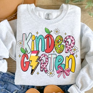 May include: A gray sweatshirt with the word "Kindergarten" written in colorful letters with a variety of patterns and designs. The letters are decorated with apples, lightning bolts, hearts, and flowers.