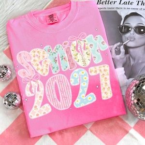 May include: A pink crew neck t-shirt with the word "SUMMIT 2021" in a patchwork design. The letters and numbers feature floral, striped, and polka dot patterns. The shirt has a Comfort Colors tag.
