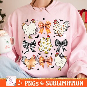 Op de afbeelding: Een roze sweatshirt met een ontwerp van kippen die verschillende kostuums dragen, waaronder een spook, een geruit overhemd en een strikje. Het ontwerp bevat ook strikken en vleermuizen.