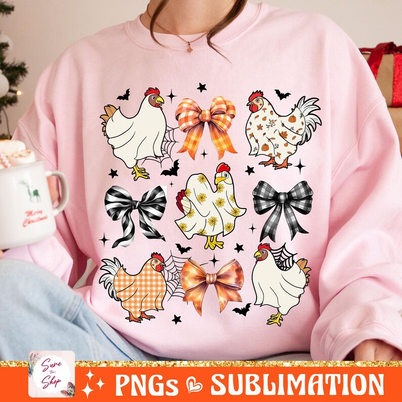 Op de afbeelding: Een roze sweatshirt met een ontwerp van kippen die verschillende kostuums dragen, waaronder een spook, een geruit overhemd en een strikje. Het ontwerp bevat ook strikken en vleermuizen.