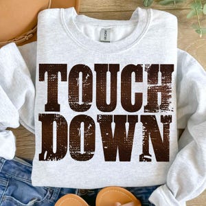 May include: A light gray sweatshirt with the words "TOUCH DOWN" printed in a distressed brown font. The sweatshirt is a crew neck style and appears to be made of a soft material. The image is taken from a top-down perspective.