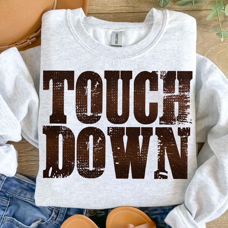 May include: A light gray sweatshirt with the words "TOUCH DOWN" printed in a distressed brown font. The sweatshirt is a crew neck style and appears to be made of a soft material. The image is taken from a top-down perspective.