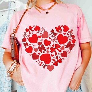 K&ouml;nnte beinhalten: Ein hellrosa T-Shirt mit einem Herzdesign aus roten Herzen und Schleifen. Das Herz befindet sich mittig auf dem Shirt, wobei unterschiedlich gro&szlig;e Herzen und Schleifen ein ansprechendes Muster bilden. Das Shirt hat kurze &Auml;rmel und ist ein l&auml;ssiges Kleidungsst&uuml;ck.