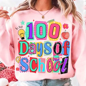 Puede incluir: Sudadera rosa claro con un gr&aacute;fico colorido que dice "100 Days of School". El dise&ntilde;o incluye una bombilla, una manzana, un l&aacute;piz y otros elementos escolares. El texto est&aacute; en varios colores y fuentes.