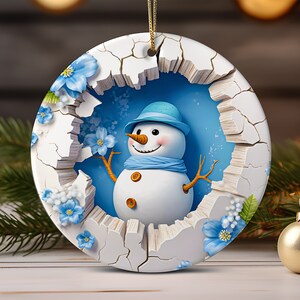 Könnte beinhalten: Ein runder weißer Keramik-Anhänger mit einer rissigen Oberfläche. Durch den Riss ist ein blauer Hintergrund mit einem Schneemann zu sehen, der einen blauen Hut und einen Schal trägt. Der Schneemann lächelt und hat eine Karottennase. Blaue Blumen befinden sich am Rand des Anhängers.