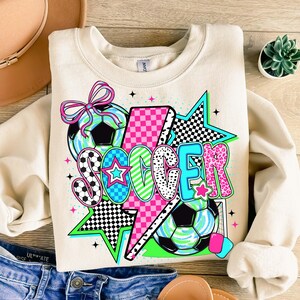 Puede incluir: Una sudadera beige con un dise&ntilde;o gr&aacute;fico de la palabra "SOCCER" en letras de colores con un rayo, estrellas y balones de f&uacute;tbol.