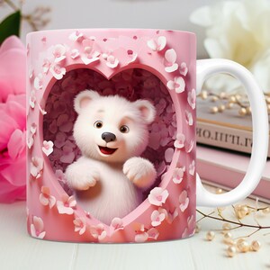 May include: A white ceramic mug featuring a smiling white teddy bear inside a pink heart-shaped frame. The frame is decorated with pink flowers. The mug has a white handle and is set against a soft-focus background.