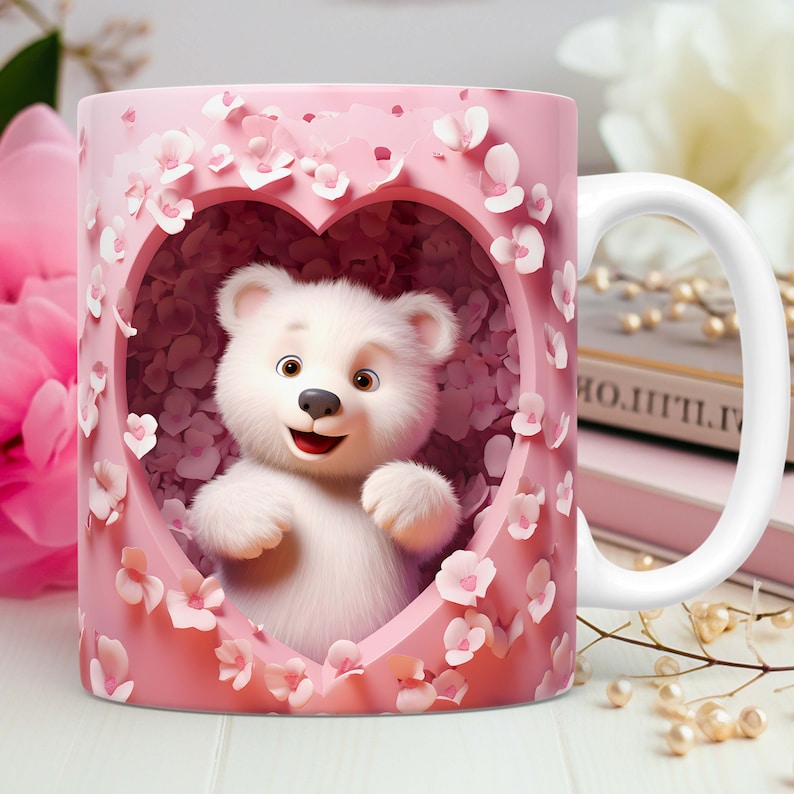 May include: A white ceramic mug featuring a smiling white teddy bear inside a pink heart-shaped frame. The frame is decorated with pink flowers. The mug has a white handle and is set against a soft-focus background.