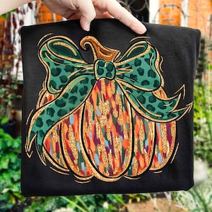 May include: Black sweatshirt featuring a colorful pumpkin design with a green leopard print bow. The pumpkin has orange, gold, and blue brushstrokes. The design is outlined in gold.