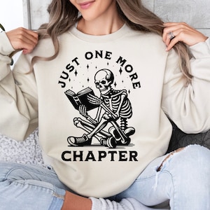 May include: A cream-colored sweatshirt with a black graphic of a skeleton reading a book. The text "Just One More Chapter" is printed around the skeleton.