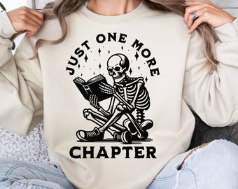 May include: A cream-colored sweatshirt with a black graphic of a skeleton reading a book. The text "Just One More Chapter" is printed around the skeleton.