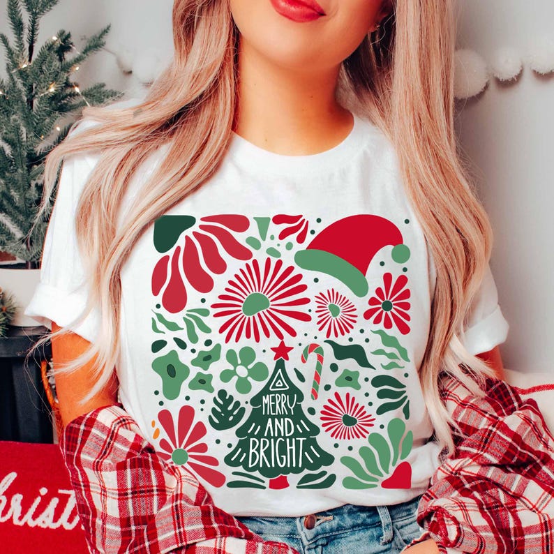 Puede incluir: Camiseta blanca con un dise&ntilde;o navide&ntilde;o festivo. El gr&aacute;fico presenta elementos florales rojos y verdes, un gorro de Pap&aacute; Noel y un &aacute;rbol de Navidad con la frase "Merry and Bright". La camiseta tiene mangas cortas.