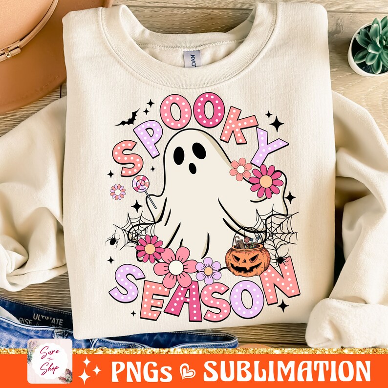 May include: A light beige sweatshirt with a pink and purple graphic design that says "Spooky Season". The design features a ghost holding a lollipop, a spider web, and flowers.