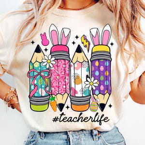 May include: White t-shirt featuring a graphic of four colourful pencils with bunny ears and the text "#teacherlife".