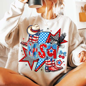 May include: A white sweatshirt with a graphic design featuring a cowboy hat, boots, and the word "USA" in red, white, and blue. The design also includes stars, checkered patterns, and a bald eagle.