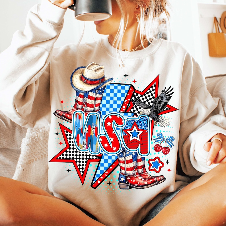 May include: A white sweatshirt with a graphic design featuring a cowboy hat, boots, and the word "USA" in red, white, and blue. The design also includes stars, checkered patterns, and a bald eagle.