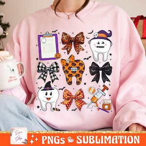 May include: A pink sweatshirt with a Halloween-themed dental design. The design features a variety of images, including teeth, bows, a clipboard, a syringe, and a mirror. The text "Sure to Shop" is visible in the lower left corner of the image.