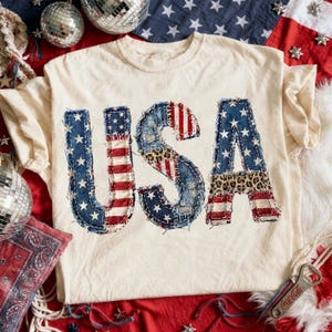 Pu&ograve; includere: T-shirt color crema con la scritta "USA" in un design patchwork. Le lettere presentano motivi in denim, stelle, strisce e stampa leopardata, evocando un tema patriottico. La maglietta &egrave; esposta su una superficie rossa e bianca con elementi decorativi.