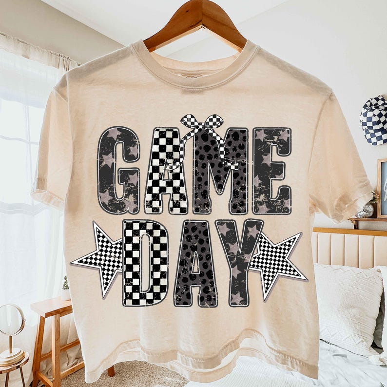 May include: Cream-colored t-shirt with the words "GAME DAY" in a distressed, checkered, and star-accented design. The shirt is hanging on a wooden hanger, suggesting a casual, sporty theme.