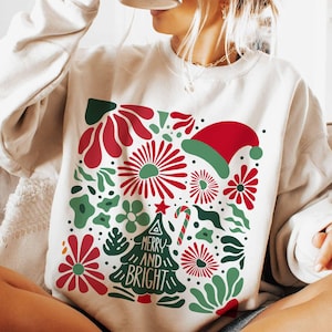 Puede incluir: Sudadera color crema con un dise&ntilde;o festivo. El gr&aacute;fico presenta elementos florales rojos y verdes, un gorro de Pap&aacute; Noel y un &aacute;rbol de Navidad con las palabras "MERRY AND BRIGHT". Un bast&oacute;n de caramelo y una estrella completan el tema navide&ntilde;o.