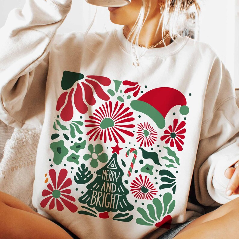 Puede incluir: Sudadera color crema con un dise&ntilde;o festivo. El gr&aacute;fico presenta elementos florales rojos y verdes, un gorro de Pap&aacute; Noel y un &aacute;rbol de Navidad con las palabras "MERRY AND BRIGHT". Un bast&oacute;n de caramelo y una estrella completan el tema navide&ntilde;o.